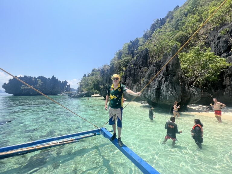 Dezza standing on the outrigger of a boat in front of limestone cliffs at Payong-Payong Beach, Miniloc Island